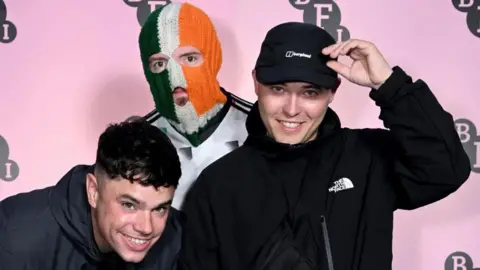 Getty Images The free members of kneecap two of who are dressed in dark clothing and the one in the middle is wearing a white sports top and Irish tricolour balaclava
