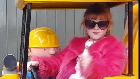 Family handout Lila is sitting in a yellow tractor and has one hand on the steering wheel. She is wearing a pink fluffy jacket and has sunglasses on.