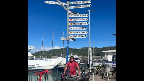 Sam Murray A man in a red t-shirt standing with his bike by a waterfront in Queensland, Australia.
