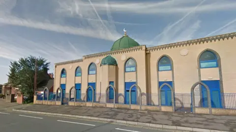 Google City Central Mosque, a brick-built building with green domed roof and religious symbols on rounded windows. There is a fence with grey brick arches along its length.