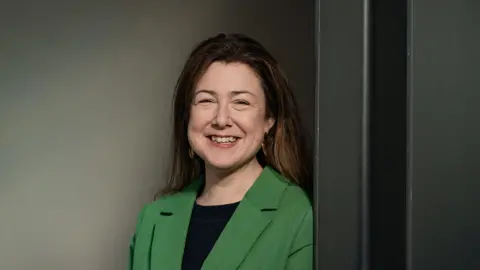 Green Party A photo of a woman with dark hair and wearing a green jacket and block top. She is stood in front of a grey coloured wall.