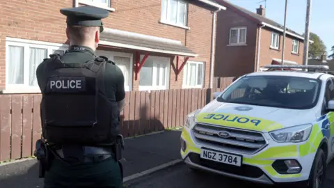 A still of the scene in Dungannon. A police officer in a green uniform and black vest with 'police' written in the back in white font stands with their back to the camera. A police vehicle is parked to the side of the officer and in the background is a red brick house.