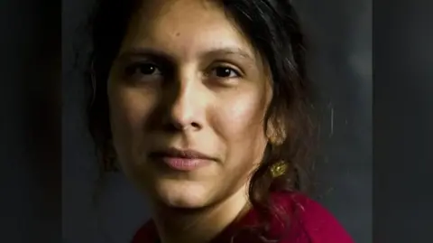 ERCC Mridul Wadhwa looking directly at the camera. She is wearing a dark coloured top and is in front of a dark background. She has a gold earring in her left ear.