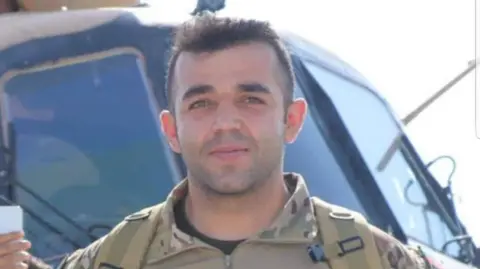 An Afghan national with short dark hair, dressed in military uniform, stood in front of a helicopter
