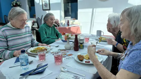 Four elderly women are sitting around a table eating fish and chips.