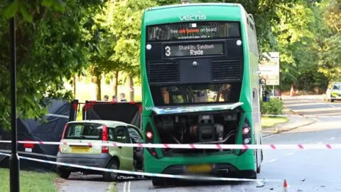 A green double-decker bus is stopped in a single-carriageway road with one lane in each direction. To the left of it, partially on the pavement, is a lime green Fiat Panda with the driver's door open. Three people can be seen behind a black tarpaulin across the pavement and adjoining grass. Police tape surrounds the scene.