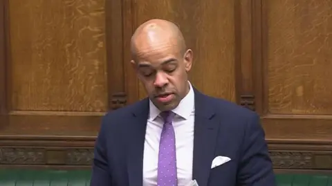 Ben Obese-Jecty stands in the House of Commons and is photographed looking down at a sheet of paper which he is reading off. He is wearing a navy suit with a purple tie. The green back benches can be seen behind him. 