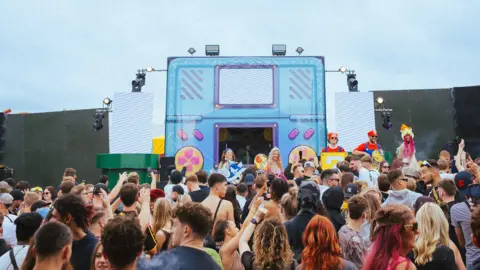 Alistair Brookes A crowd is seen from behind, looking at a stage that resembles a Gameboy. It has a canvas depicting a small screen and buttons. There are several people dressed like Super Mario characters facing the crowd.