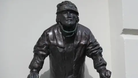 A bronze statue of Brian Haw wearing a chapeau  and jacket.