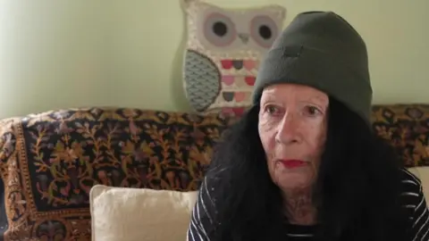 Sharon Lawson, a woman with long black hair wearing a grey beanie hat and black and white striped top, sits on a patterned sofa in her flat