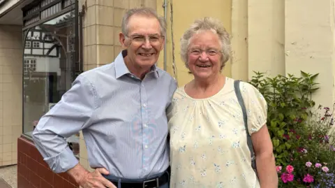 An older couple stand shoulder to shoulder as they face the camera and smile. The man is wearing a light blue long-sleeved shirt and the woman has an open-necked light yellow top with white patterning on it
