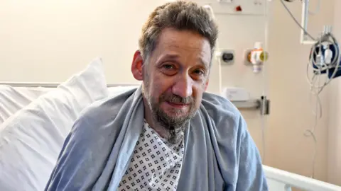 Royal Papworth Hospital Daniel Evans-Smith looks at the camera from his hospital bed. He is dressed in a hospital gown with a blanket over his shoulders. He had a beard and greying hair. You can see oxygen pipes in the background which are not attached to Mr Evans-Smith