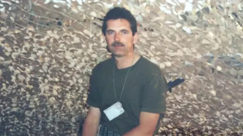 Mike Marshall Mike Marshall while serving in the army. He is sitting down with a camouflage netting above him. He is wearing a green khaki T-shirt is looking directly at the camera. He has dark hair and a moustache and is wearing a chain round his neck.