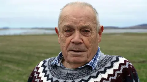 David is looking into the camera. He is wearing a patterned jumper. He is standing in an area of coastal meadow called machair.