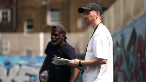 Michael Johnstone stands holding papers in his hands and wears a whistle and a cap. A man stands behind him.