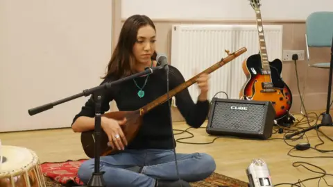 Confluence Collective Oxford Della Izadi sitting cross legged in a room, playing a string instrument. There are a guitar, a drum and microphones around her as well. She is not looking at the camera.