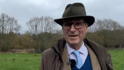 Robby West/BBC Henry Cator, a man who is standing in a field in Taverham. He is looking directly at the camera and smiling and is wearing a shirt with a pale blue tie, with a blue jumper over the top and two brown coats. He is wearing a hat on his head.