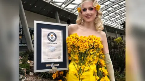 Marmalade wearing a yellow dress covered in daffodils. She is holding a Guinness World Records certificate. 