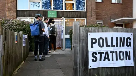 Getty Images A group of people stand in a queue outside a community centre - the sign outside pinned to a fence indicates it is a polling station. 