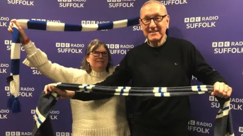 A woman and man hold football scarves and wave them in the area. The woman has short dark grey hair, glasses and is wearing a cream knitted jumper. The man is bald, wearing glasses and a black jumper.