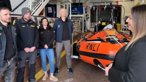 A woman speaks to three men and a woman in a lifeboat shed