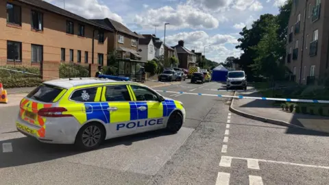 A police car in a residential street in Kings Langley, further police activity can be seen in the distance, including a police tent 