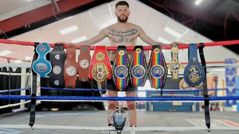 Jason Cunningham is wearing dark shorts and white boots. He has short brown hair, a brown beard and tattoos across his chest. He stands inside a boxing ring behind a row of championship belts draped across the top rope. A large silver trophy is positioned on the ring floor in front.
