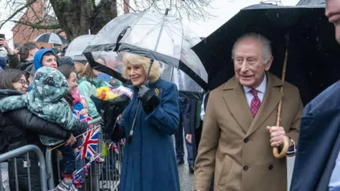 PA Media The King and Queen meeting the public. Both of them are holding umbrellas and wearing coats. There are metal gates lining the road, and crowds behind them in waterproof clothing.
