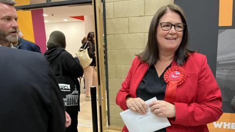 A smiling Rachel Hopkins looking to the left of the camera, dressed in a red suit jacket with a red Labour rosette on the lapel. She is clutching a white envelope and two people stand to her right. 
