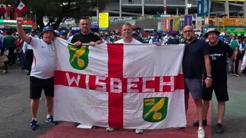 Five men holding up a St George cross flag with 'Wisbech' written on it and the emblem of Norwich City FC too. Paul Green is on the far left with a white England football shirt and black knee length shorts.