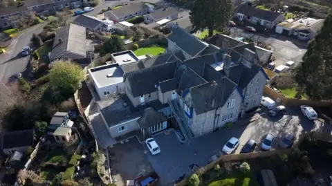 Aerial view of the former St Monica’s Maternity Home, now a large multi-section building with pitched roofs and several extensions, surrounded by a residential neighbourhood of bungalows, parked cars and small gardens.
