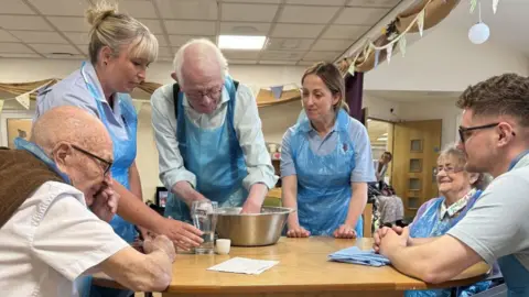 RBL Natalie is standing at a table with a blue apron on, watching an older man with white hair and black glasses knead dough in a silver bowl. A woman is standing beside him and an older man and woman are sitting at the table near them, also wearing blue aprons. A younger man also sits at the table