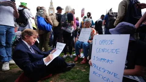 PA Media A man in a suit writes on a placard, while others sit near him holding signs
