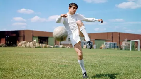 PA Media Norman Hunter in a full white football kit. He is mid air about to kick a football on a green field.
