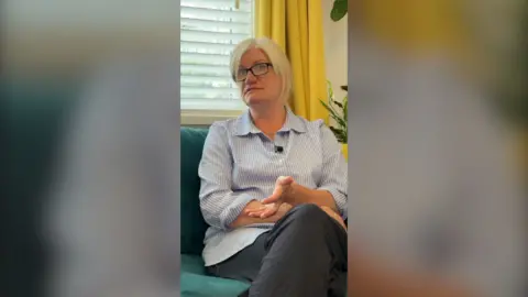 Blonde woman wearing a light blue stripped shirt sits on sofa during interview