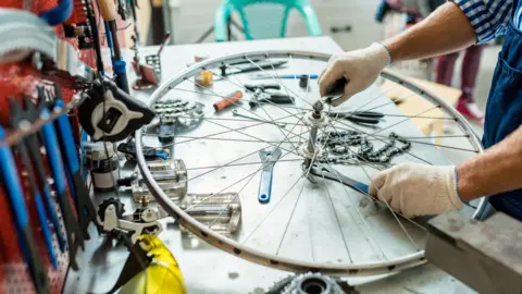 A bike mechanic works on a bicycle wheel on a worktop. There are an assortment of tools on the table and on a wall next to it.