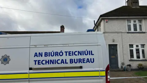 A white house, taped off with police tape
There is a white Garda van that reads technical bureau parked outside