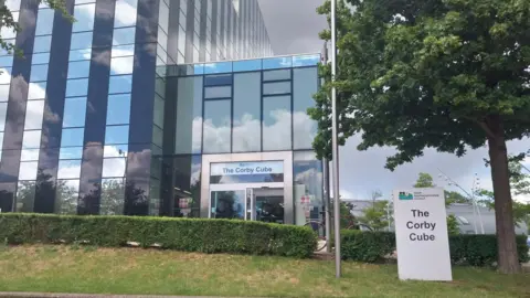 LDRS/PA Media The exterior of the Corby Cube, with two signs showing that name. It is a tall modern building with reflective windows.