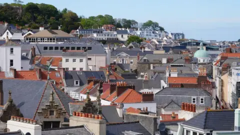 Am image of houses and red and grey rooftops in Guernsey, with trees in the background. The sky is blue.
