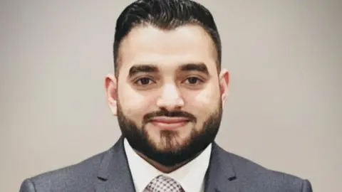 Umer Ali A man in a grey suit with short black hair and beard