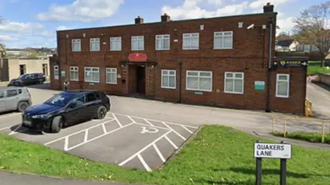 A two-storey brick built building stands surrounded by a car park and grassy area.