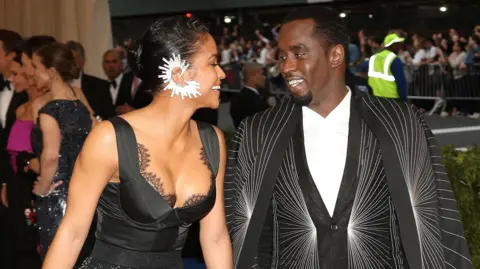 EPA Cassie Ventura and Sean Combs smile at each other at the 2017 Met Gala. She wears a black dress with a large white earring. He wears a white shirt and a black and silver Rick Owens suit and matching cape decorated with a striking spider-web-like pattern. 