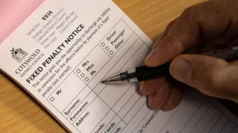 A close-up of a hand holding a pen filling out a fixed penalty notice from Cotswold District Council