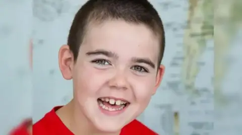 CLC Portraits A boy with dark hair and his mouth open is looking at the camera. He is wearing a red top.