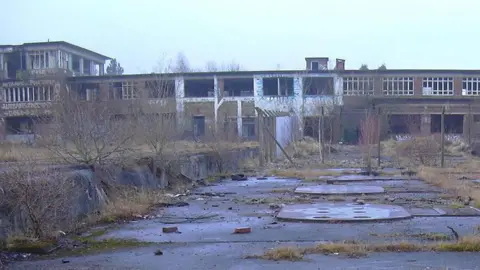 The deserted site of Huncoat power station. It is a long two-storey brick building with a taller section to the left which have been vandalised. To the front are overgrown flat areas with covers on the ground and sprouting weeds