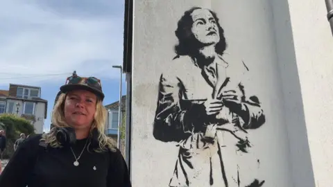 A young woman stands next to the mural on the steep hill. She wears a cap and has headphones around her neck.