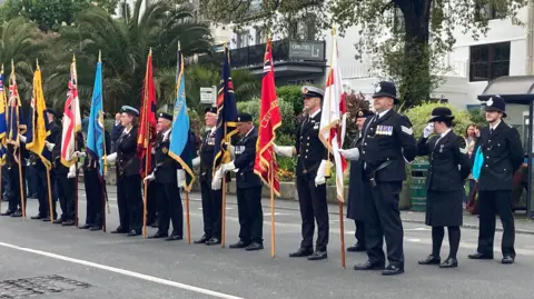 A line of police officers and military personnel stand on a road holding flags during Guernsey's Liberation Day in 2023. They are stood in front of a white building with several trees and bushes.