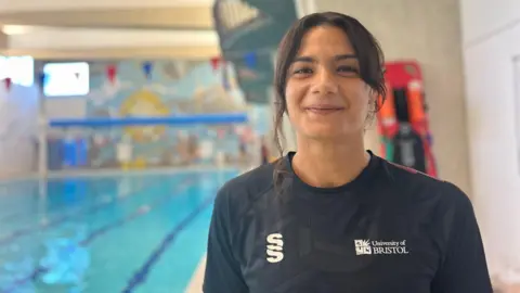 Bristol University and schools swimming teacher Amaya Scott with dark hair and a navy blue t-shirt with a the University of Bristol logo on it smiling and standing in front of a blurred background of an indoor swimming pool.