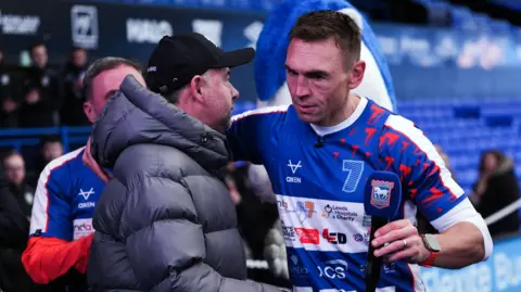 PA Media Marcus Stewart and Kevin Sinfield embrace in a hug inside Portman Road stadium. Stewart wears a black cap and grey puffer coat. Sinfield wears a blue running top with a white long sleeved top underneath. He holds a microphone in his left hand which has the Ipswich Town Football Club logo on it. 
