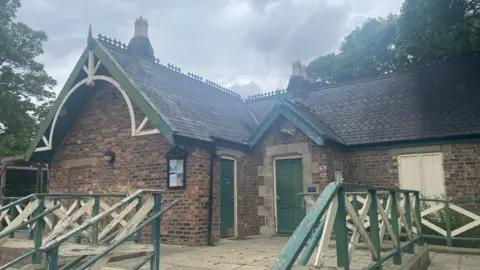 A red brick, single-storey Victorian pavilion with a slanted roof with green wooden trim around it. There are two green doors on the right of the building and a green and cream wooden banister around the path in front of it. The paintwork is chipped, making the building and banisters look run down. There is a bulletins board at the front of the building.